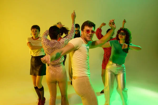 Sarı tişörtlü gülümseyen adam neon altında retro dans grubunu yönetiyor. Bireysellik kavramı, gece hayatı performansı, kültürel kimlik, neşeli müzik ve eğlence tarzı.