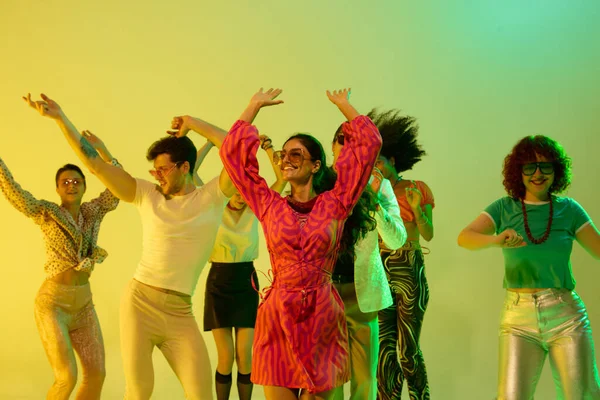 Pembe elbiseli gülümseyen kadın grupla neon sarısı altında dans ediyor. Neşeli gece hayatı, retro moda, kutlama, kültürel birlik ve enerjik müzik tarzı kavramı.
