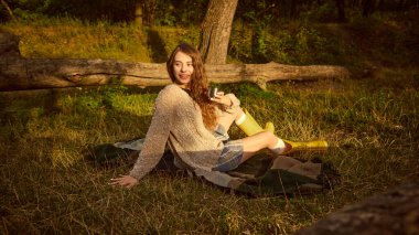 Ormanda battaniyeye oturmuş altın ışıkta sıcacık gülümseyen genç bir kadın. Açık hava yaşam tarzı, rahatlama, sonbahar modası, rahat yaşam ve neşeli duygu kavramı..