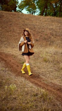 Sarı çizme, sonbahar ceketi ve şapkasıyla yokuş aşağı yürüyen şık bir kadın. Sonbahar modası, açık hava yaşam tarzı, kırsal eğlence, mevsimlik yürüyüş ve kendine güvenen ruh hali kavramı..