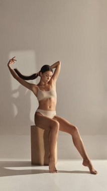 Bej iç çamaşırlı, kollarını esneten ve zarif bir şekilde tahta küpte poz veren kadın model. İç çamaşırı modası, editör fotoğrafçılığı, kendine güven ve kadın tarzı kavramı..