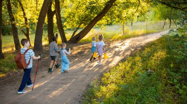 Bir grup çocuk ağaçların arasından gün ışığıyla orman yolunda yürüyorlar. Aile yaşam tarzı kavramı, açık hava macerası, çocukluk keşfi, eko-eğitim ve doğa bağı..