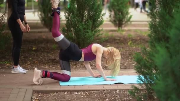 Athlète femme impliquée dans la remise en forme dans le parc, faire des levées de jambes sur un tapis avec entraîneur — Video