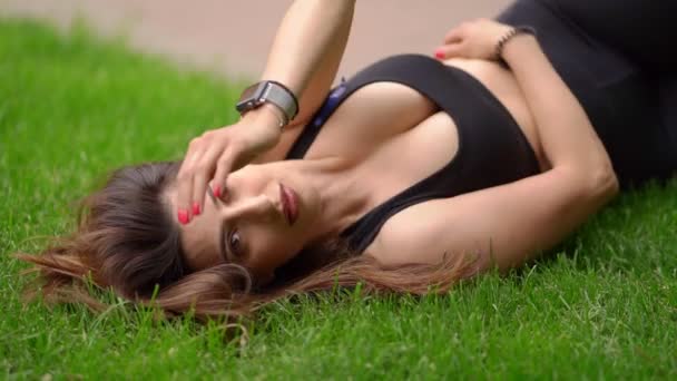 Atleta chica con hermosos pechos está descansando en el césped en el parque después de un entrenamiento . — Vídeos de Stock