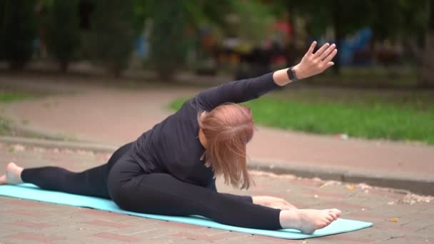 Dívka sportovec ve sportovním oblečení strečink, sedí na štěpení v parku. — Stock video