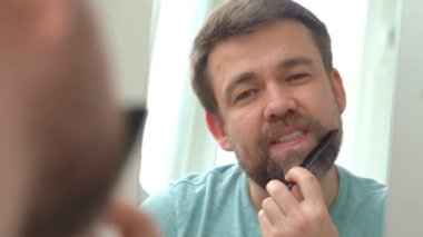 Tişörtlü bir adam evde aynanın önünde sakalını tarıyor..