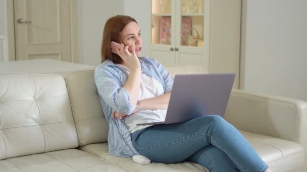 Kobieta siedzi na kanapie z laptopem i rozmawia przez telefon. dzieli się uczuciami z przyjacielem. — Wideo stockowe