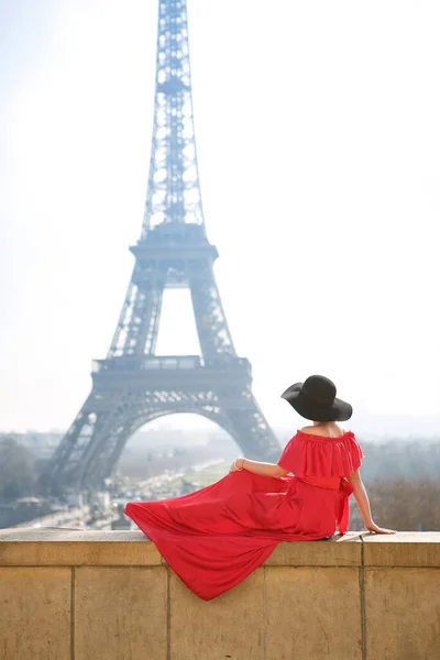Nő piros ruhában és kalapban ül egyedül előtt Eiffel torony Párizsban. — Stock Fotó