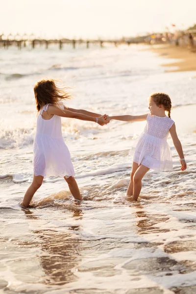 Meisje in witte jurken spelen op het strand. oudere zus trekt jonger in het water. — Stockfoto