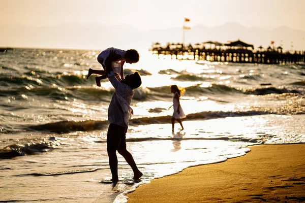 Sylwetka. Tata z dziećmi spacerujący wzdłuż brzegu morza w wietrzną pogodę. zabawna rodzina. — Zdjęcie stockowe