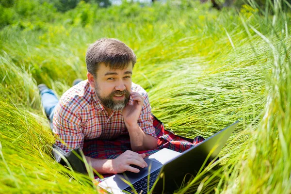 El hombre aprende, trabajando en el portátil que yace en el prado. Internet móvil en el medio rural . — Foto de Stock