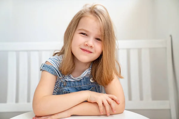 穿着粗斜纹棉布的小女孩坐在桌旁的白色长椅上。儿童明亮的工作场所 — 图库照片
