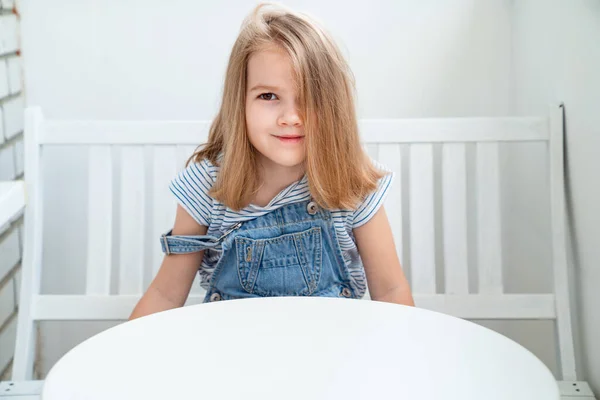 Klein meisje in denim zit op een witte bank aan tafel. lichte werkplek voor kinderen — Stockfoto