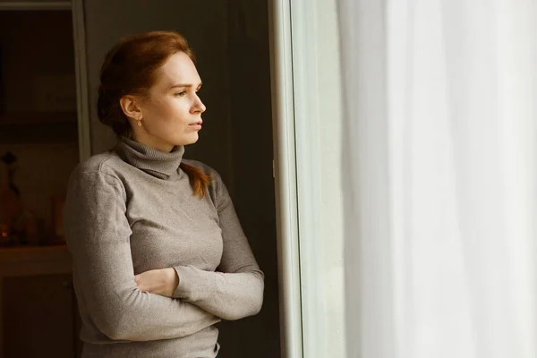 sad girl standing at window and looks into distance. upset woman waiting someone