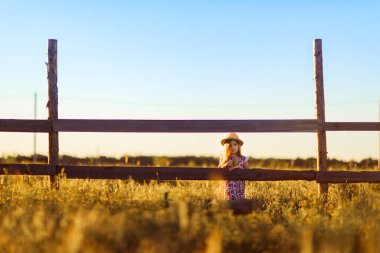 Gün batımında şapkalı kız köyde çitin yanında duruyor. Kırsalda yürür.