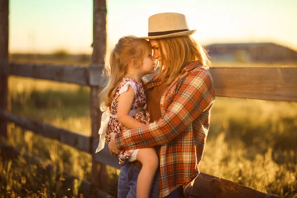 Anne ve kızı şapkalı, ekose gömlekli köyde gün batımında çitin yanında duruyor.