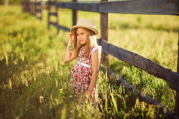 Niña, niña con sombrero de pie junto a la valla en la aldea. paseos por el campo . — Foto de Stock