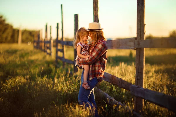 Madre e hija en sombrero, camisa a cuadros de pie cerca de la cerca al atardecer en la aldea —  Fotos de Stock