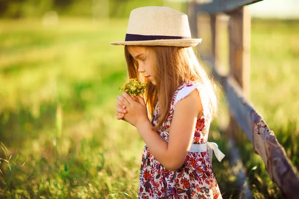 Chica en sombrero de pie junto a la cerca en la aldea con ramo de flores silvestres —  Fotos de Stock