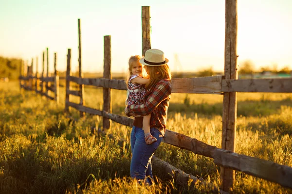 Moeder en dochter in hoed, ruitvormig hemd bij zonsondergang in dorp — Stockfoto