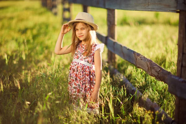 Niña, niña con sombrero de pie junto a la valla en la aldea. paseos por el campo . — Foto de Stock