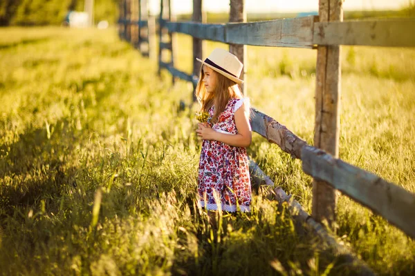 Mädchen mit Hut steht neben Zaun im Dorf mit Strauß Wildblumen — Stockfoto