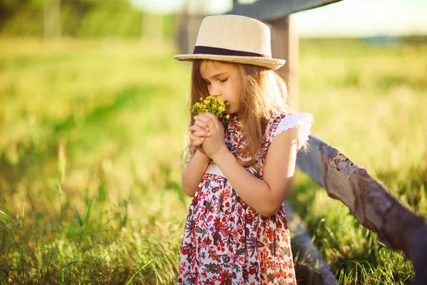 Chica en sombrero de pie junto a la cerca en la aldea con ramo de flores silvestres —  Fotos de Stock