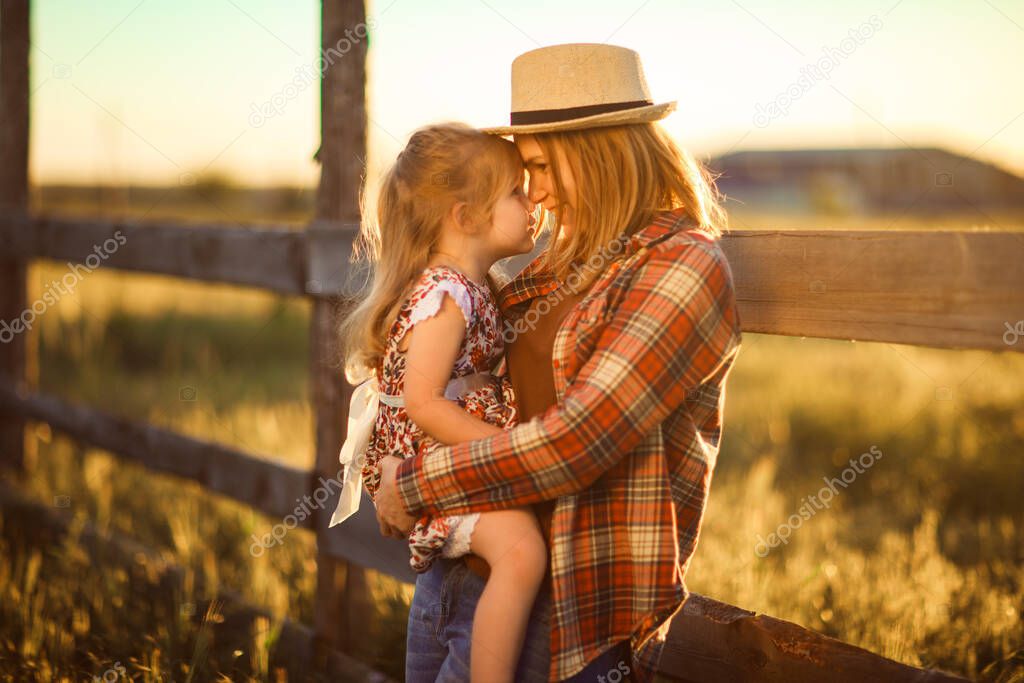 mother and daughter in hat, plaid shirt standing near fence at sunset in village