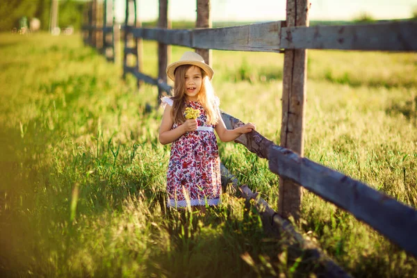 Girl in hat standing next to fence in village with bouquet of wild flowers — Stock Photo, Image