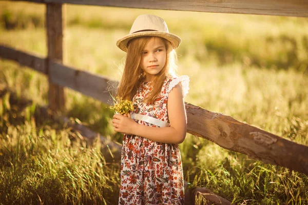 Girl in hat standing next to fence in village with bouquet of wild flowers — Stock Photo, Image