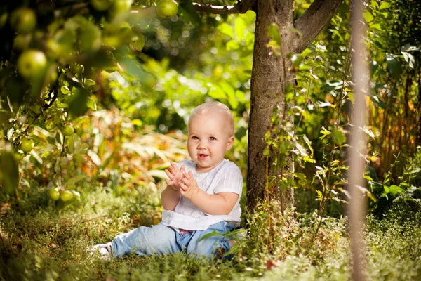 Liten flicka sitter på gräs i trädgården under små äppelträd. Trädgårdsodling. — Stockfoto