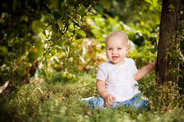 Liten flicka sitter på gräs i trädgården under små äppelträd. Trädgårdsodling. — Stockfoto