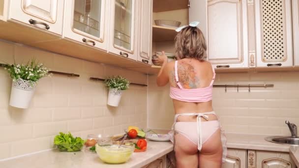 Sexy huisvrouw blond in slipje en schort op zoek naar gerechten in keuken. — Stockvideo