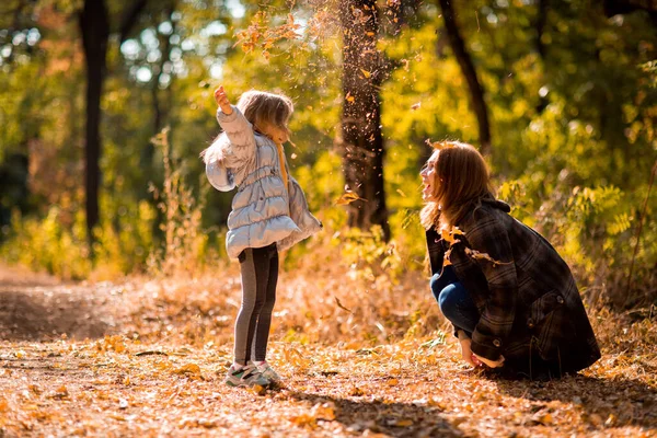 Мама играет с дочерью, бросает сухие желтые опавшие листья. прогулки в осеннем парке . — стоковое фото