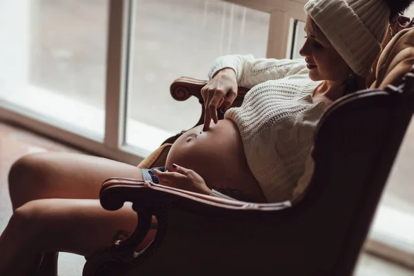 Gravid flicka med tatuering i stickad mössa sittande i fåtölj framför fönstret. — Stockfoto