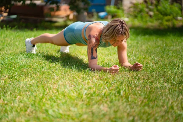 锻炼板，手倒立。有纹身的金发女孩在花园里做运动. — 图库照片