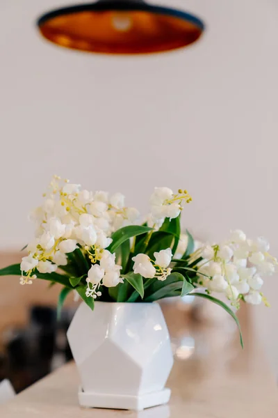 Vas putih kecil dengan bunga lili lembah di atas meja di bawah lampu gantung tembaga hitam. — Stok Foto