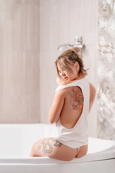 Sexy Mädchen mit Tätowierungen in weißem Tank-Top sitzt am Rand der Wanne. Hautpflege. — Stockfoto
