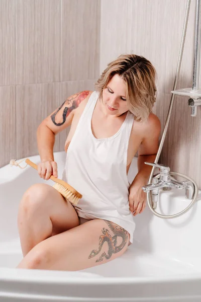 Сексуальная девушка с татуировками делает целлюлитный массаж специальной щеткой в ванной комнате . — стоковое фото