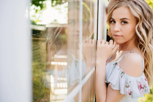 Hermosa chica rubia en vestido de verano de pie junto a la ventana en el jardín en el patio trasero . —  Fotos de Stock