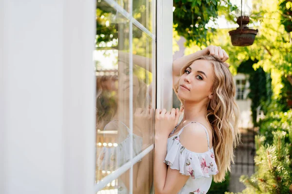 Yazlık elbiseli güzel sarışın kız bahçede pencerenin önünde duruyor.. — Stok fotoğraf