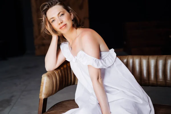 Hermosa rubia en vestido blanco sentado en el sofá de cuero en el loft . — Foto de Stock