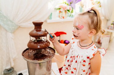 Yüz boyalı kız fıskiyeden çikolata yiyor.