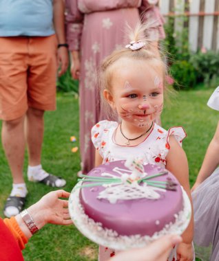 Çimlerde doğum günü pastası yiyen yüzü boyalı bir kız..