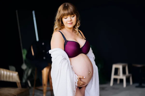Gravid kvinna med övervikt i underkläder och vit mantel stroke mage. — Stockfoto