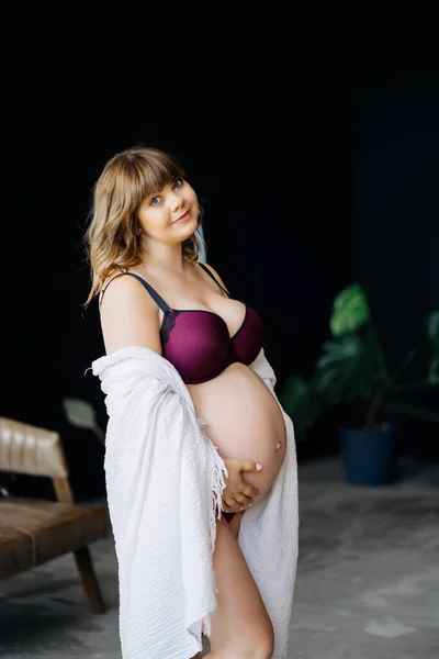 Femme enceinte en surpoids en sous-vêtements et manteau blanc caressant le ventre. — Photo