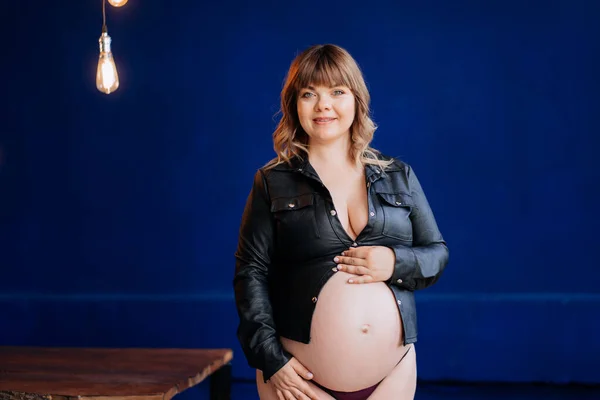 Topless. femme enceinte nue dans une chemise en cuir posant contre un mur bleu. — Photo