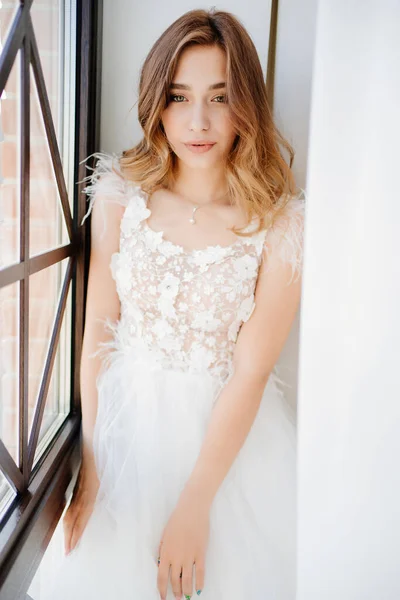 Hermosa novia en vestido blanco se sienta por la ventana. — Foto de Stock