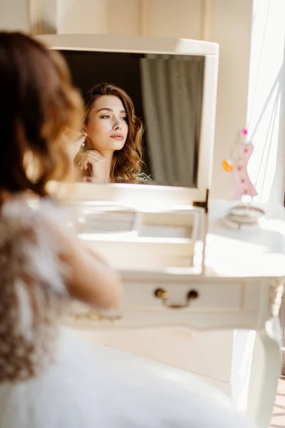 Bride is sitting near mirror and wears earrings. — Stock Photo, Image