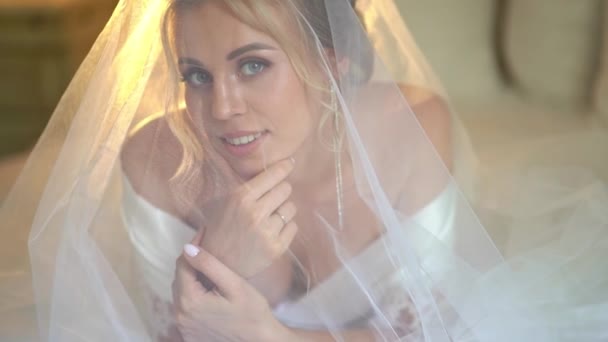 Bela noiva loira sob véu e em um manto de seda deitado na cama . — Vídeo de Stock